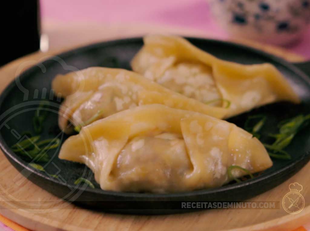 Três guiozas cozidos e macios, com um leve dourado na base e massa clara e úmida por cima, servidos em uma pequena frigideira de ferro fundido. Eles estão guarnecidos com cebolinha picada e apoiados sobre uma tábua de madeira.